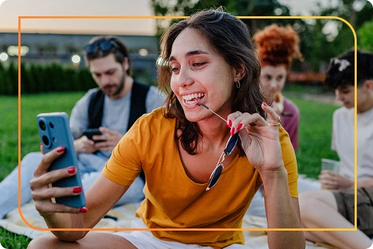 Woman in an orange top smiling and looking at her phone, sitting outdoors with friends in the background, framed by a yellow-orange rectangle graphic.